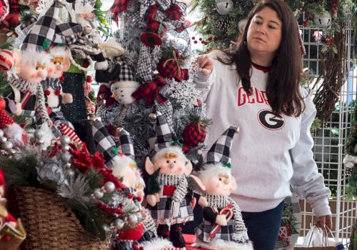 A woman in a white sweater with a shopping bag admires various holiday decorations, including elves and a snowman, displayed on a festive table.