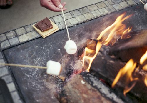 People are roasting marshmallows over a fire pit, preparing ingredients for s'mores with chocolate pieces and graham crackers nearby.