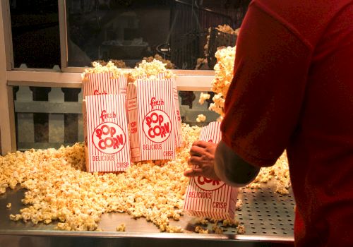 A person filling striped bags with popcorn at a counter, with popcorn scattered around. The bags are labeled 