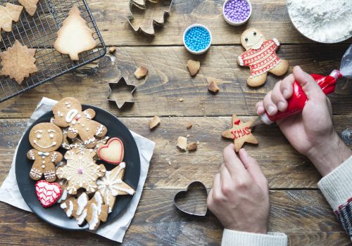 A person decorates a star-shaped cookie with icing. Various Christmas-themed cookies and baking supplies are scattered on a wooden table.