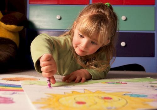 A child is lying on the floor, drawing with crayons on a large sheet of paper, with a colorful drawer set in the background.