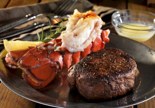 A plate featuring a juicy steak and a buttered lobster tail, garnished with a lemon wedge and rosemary, with a small bowl of melted butter on the side.