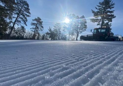 A snowy landscape with trees in the background and a vehicle, possibly a snow groomer, creating neat lines in the snow under a bright sun.