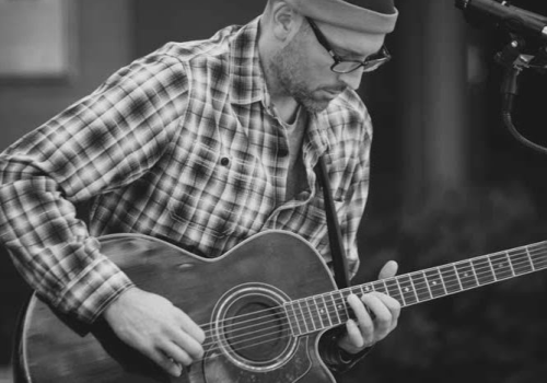 A bearded individual wearing glasses and a beanie plays an acoustic guitar while standing near a microphone, captured in a black-and-white photo.