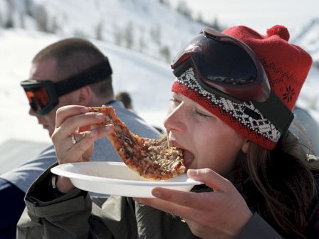 A person wearing a red beanie and ski goggles is eating a slice of pizza while another person with ski goggles is in the background in a snowy setting.
