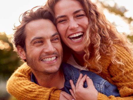 A happy couple, with the woman in a yellow sweater, smiling and embracing outdoors. The background has a blurred, sunny setting with greenery.