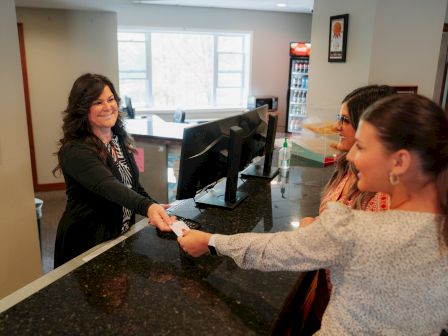 A receptionist hands a keycard to two women at a front desk, with a computer and office supplies visible in the background.
