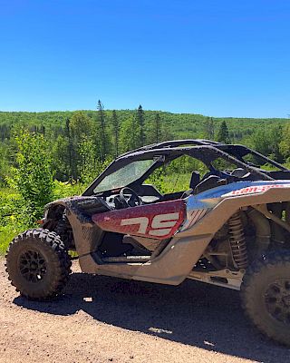 An off-road vehicle is parked on a dirt path with a lush, green forest and clear blue sky visible in the background.